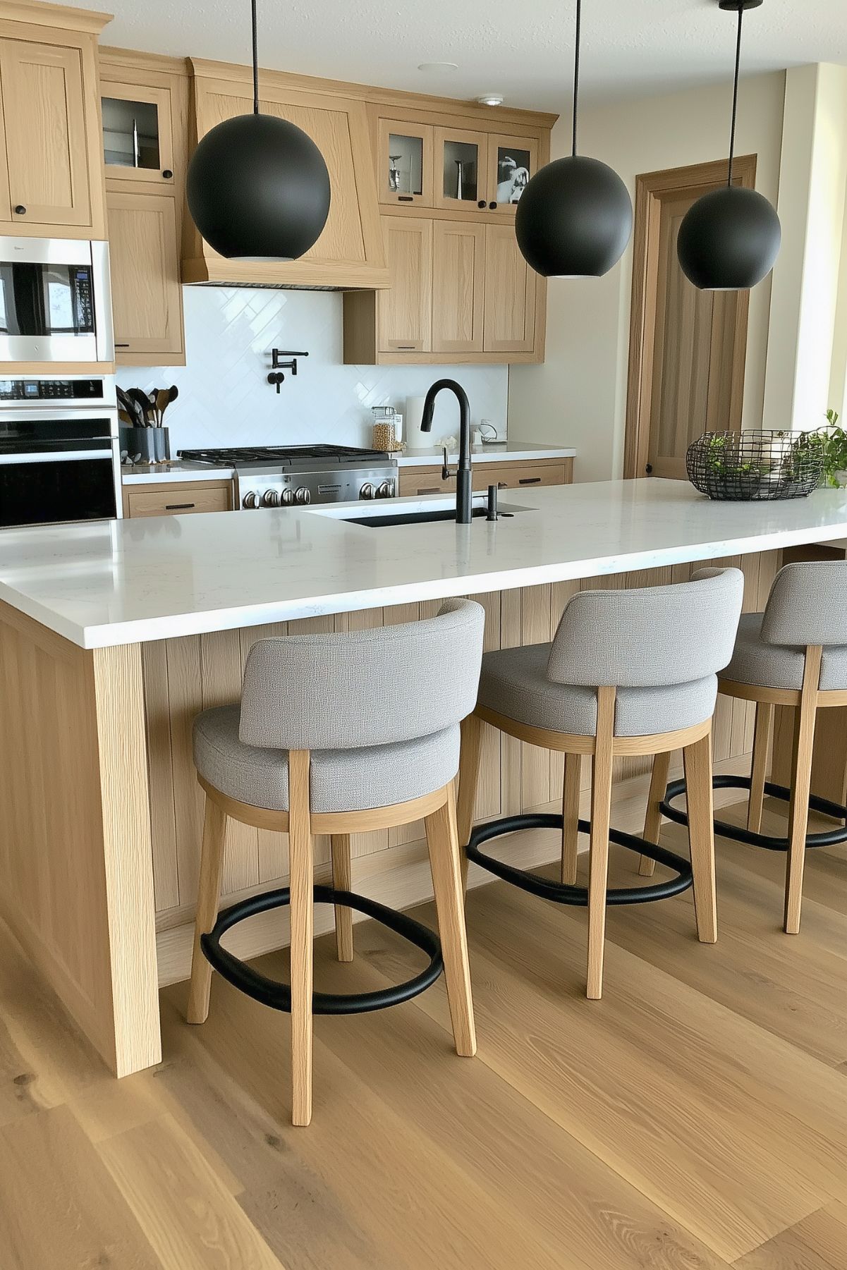 Modern kitchen with white oak cabinetry and a matching island featuring a sleek white quartz countertop. Three matte black spherical pendant lights hang above the island, complementing the black faucet and hardware. Light gray upholstered bar stools with wooden legs and black footrests provide seating. The backsplash features a subtle white herringbone pattern, adding texture without overpowering the space.