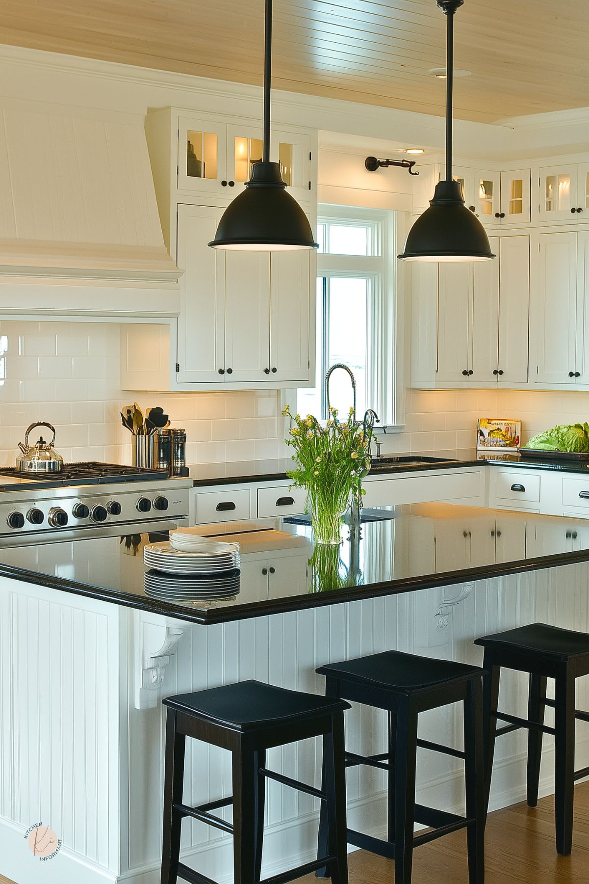White beadboard island with a polished black granite waterfall countertop and three black wood stools beneath matte black pendant lights. Cream shaker cabinets with black cup pulls and a subway tile backsplash frame a stainless steel range. A chrome spring-neck faucet, bouquet of wildflowers, and black hardware add modern contrast in this chic kitchen design.