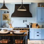 Matte black custom range hood and matching pendants hover over a black-and-wood island with leather-cushioned black spindle stools. Powder blue shaker cabinets with black cup pulls flank a marble-topped counter and stainless range with gold knobs. White square tile backsplash, brass pot filler, a wood tabletop island and potted branches complete this modern dramatic kitchen design.