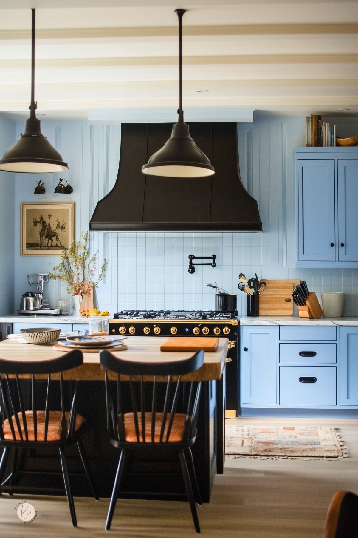 Matte black custom range hood and matching pendants hover over a black-and-wood island with leather-cushioned black spindle stools. Powder blue shaker cabinets with black cup pulls flank a marble-topped counter and stainless range with gold knobs. White square tile backsplash, brass pot filler, a wood tabletop island and potted branches complete this modern dramatic kitchen design.