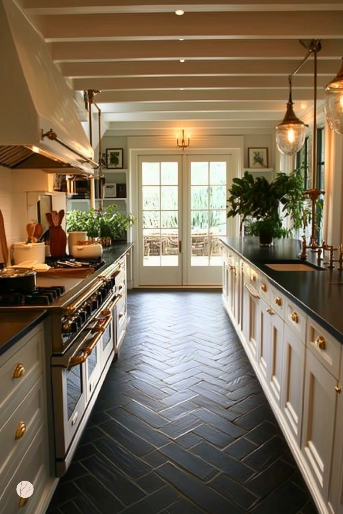White shaker cabinets with brass cup pulls flank a black quartz countertop and inset farmhouse sink under a custom white hood. Matte black subway tile floor in a herringbone pattern leads to glass French doors. Exposed white ceiling beams, marble-topped islands with brass pot fillers, potted herbs and copper pendants add warmth and dramatic contrast.