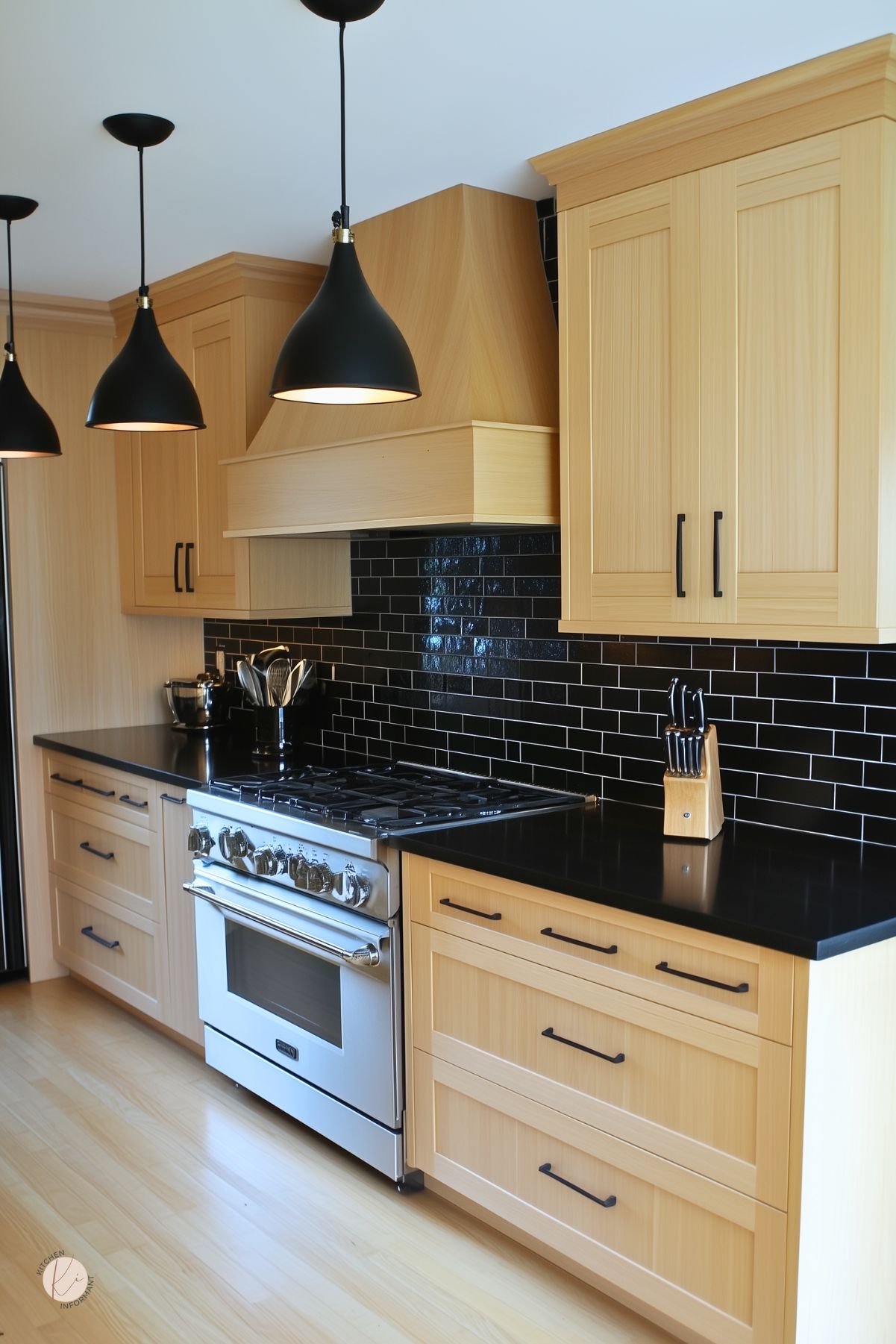 Light oak cabinets with black bar pulls and black quartz countertops set against glossy black subway tile. Three matte black pendant lights illuminate a stainless steel gas range under a wood-clad vent hood. Light hardwood floors complete a modern kitchen design with chic black accents.