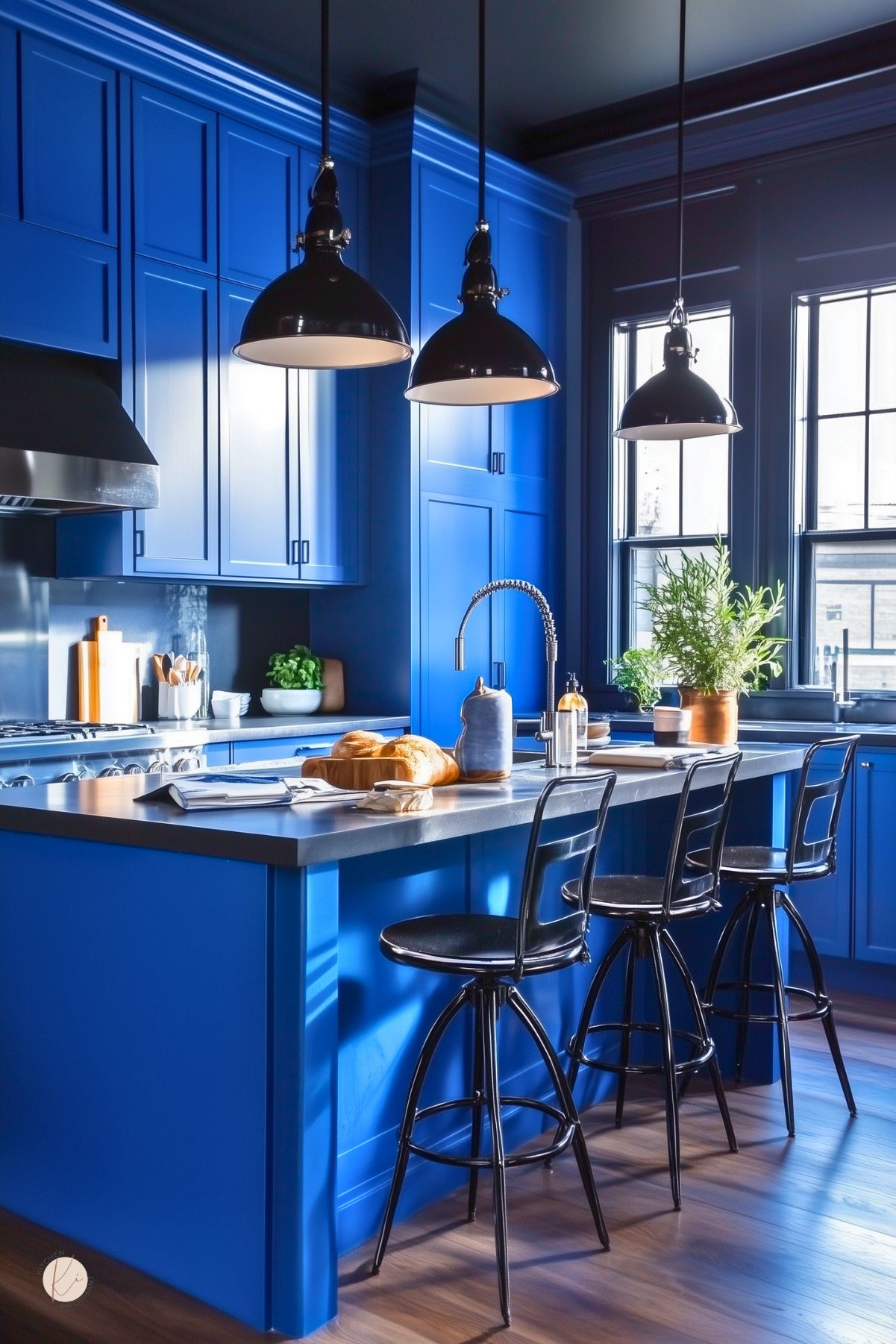 Cobalt blue floor-to-ceiling shaker cabinets with black hardware frame a gray quartz island topped by a stainless steel spring-neck faucet. Three glossy black industrial pendants hang over black metal stools at the blue kitchen island, styled with a loaf of bread, open notebook, and potted herb for a modern dramatic kitchen design.
