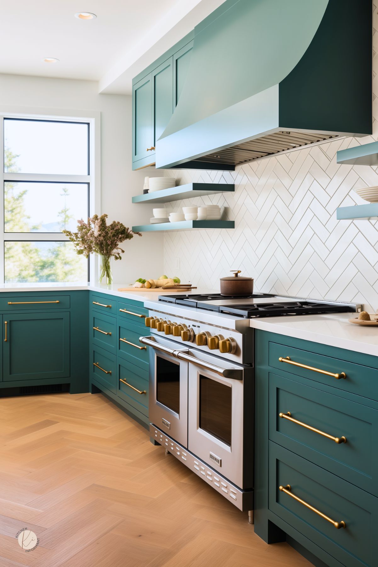Stainless steel double-oven range with brass knobs sits beneath a custom teal vent hood. White herringbone tile backsplash and matching teal open shelves display white dishes. Teal shaker base cabinets with brass bar pulls flank the range under a white quartz countertop that holds a clear glass vase of dried blooms. Light wood herringbone floors run beneath.