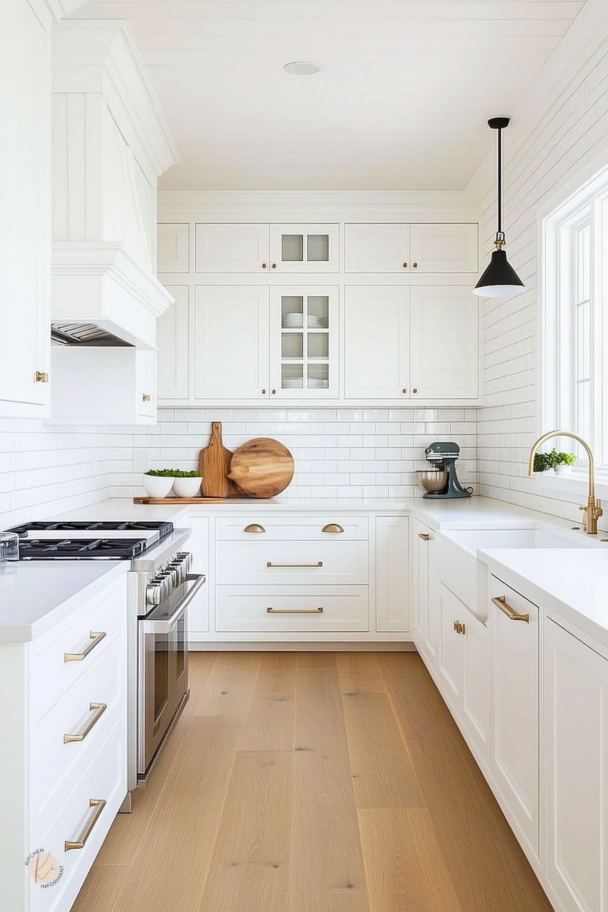 Bright white kitchen with shaker cabinets, subway tile backsplash, and brass hardware. A farmhouse sink with a brass faucet sits beneath a window, while wood cutting boards and bowls add warmth. Light wood flooring enhances the clean look, complemented by a black pendant light and a stainless steel range for a timeless finish.