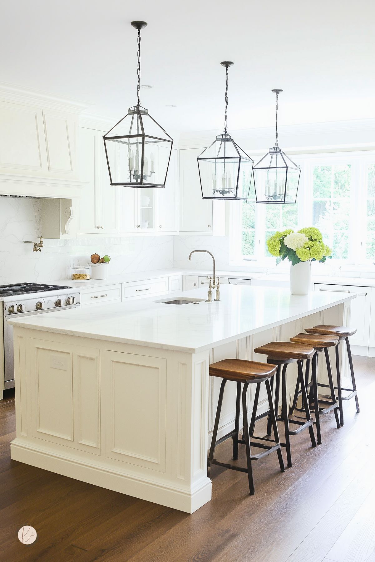 Elegant white kitchen with a large marble island and paneled cabinetry. Black metal lantern pendant lights hang above, while wooden stools with black bases provide seating. White quartz countertops and marble backsplash create a seamless look, complemented by brass fixtures. A vase of green hydrangeas adds a fresh touch near the bright windowed wall.
