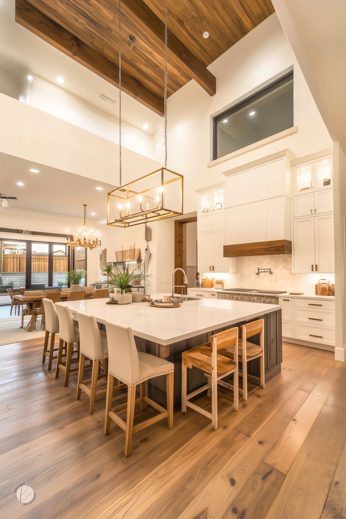 Spacious kitchen with a soaring ceiling and exposed wood beams, featuring a large marble island with seating for seven. White shaker cabinets and a custom range hood with wood trim pair beautifully with a herringbone backsplash. A brass rectangular chandelier hangs above the island, while warm wood floors and natural light create a welcoming, timeless feel.