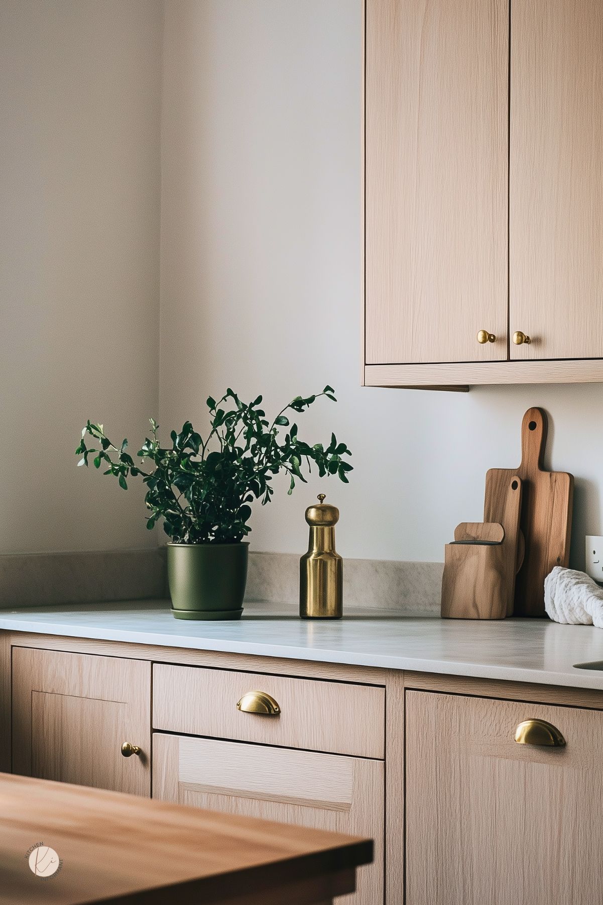 Minimal cozy kitchen with light oak cabinets, white countertop, and brass hardware. A green potted plant and brass mill sit beside stacked wooden cutting boards; soft beige walls and simple styling. Warm neutral kitchen design, Scandinavian vibe. Text: “Kitchen Informant.”