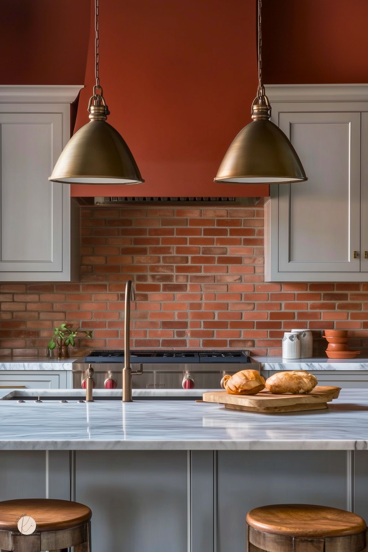 Cozy kitchen with terracotta walls and matching range hood, brick backsplash, and light gray shaker cabinets. Two large brass pendants hang over a marble island with a brass faucet, wood stools, and loaves of bread on cutting boards. Stainless range with red knobs. Warm rustic-modern kitchen design. Text: “Kitchen Informant.”
