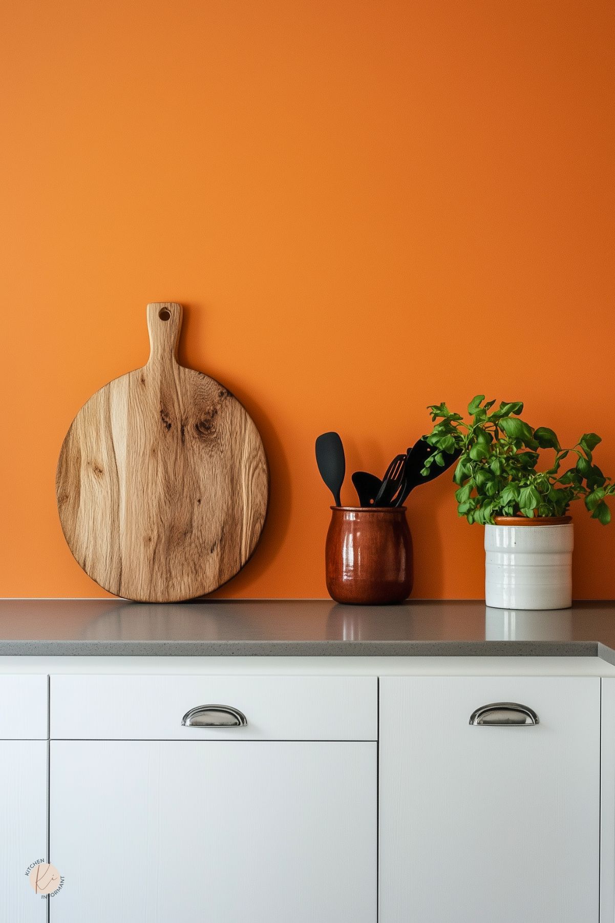 Bright cozy kitchen vignette with a bold orange accent wall, white lower cabinets with cup pulls, and a gray countertop. Round wood cutting board leans against the wall beside a brown crock of black utensils and a white pot with fresh basil. Minimal modern kitchen decor. Text: “Kitchen Informant.”
