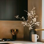 Moody modern kitchen with matte black cabinets, brass hardware and faucet, and a light quartz countertop. Warm taupe plaster walls, gas cooktop, and a black vase of white blossoms beside a stoneware mug. Soft window light creates cozy ambiance. Text: “Kitchen Informant.”