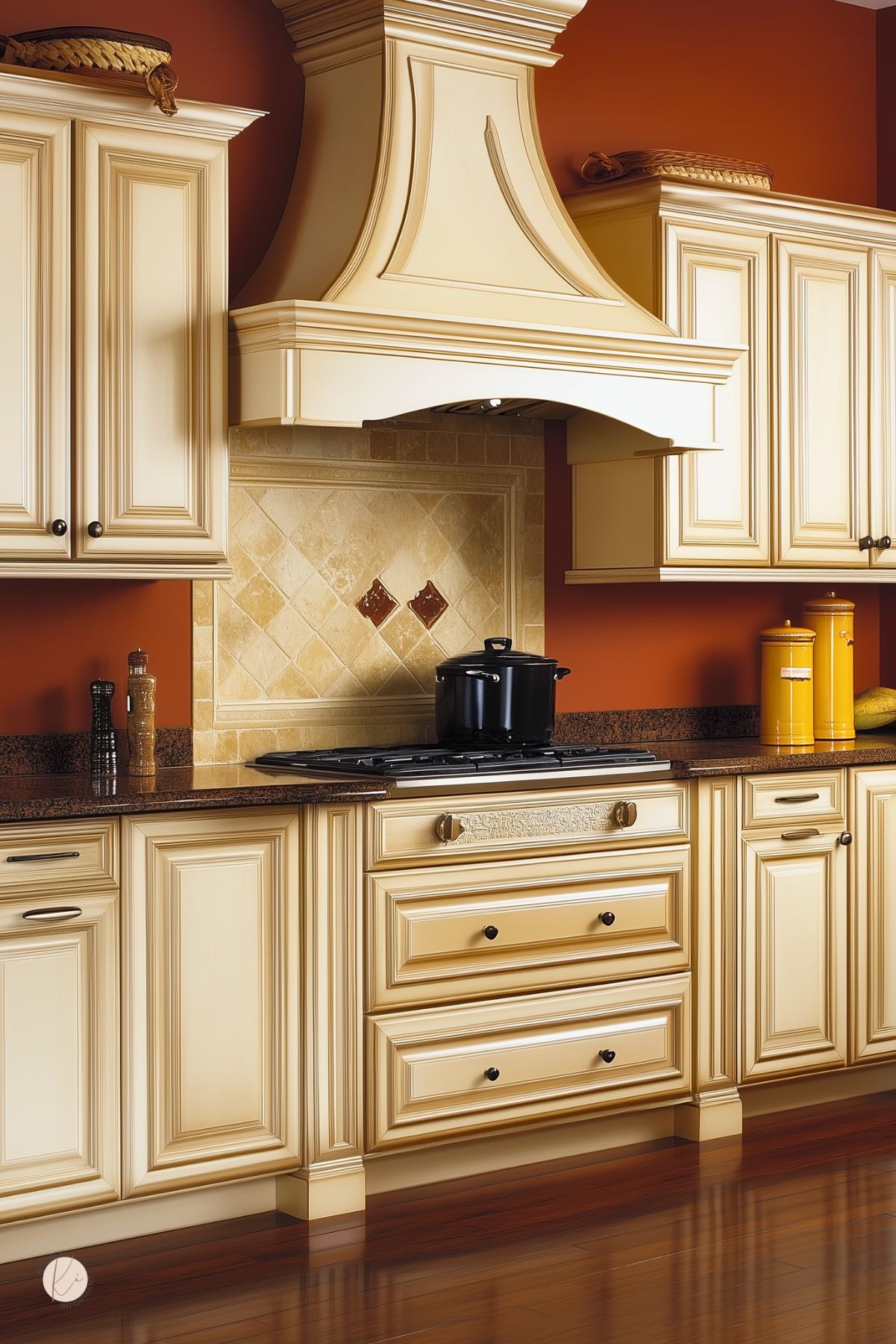 Traditional cozy kitchen with terracotta walls, cream raised-panel cabinets, and an ornate range hood. Tumbled stone tile backsplash with diamond accents, dark granite countertops, gas cooktop with black Dutch oven, yellow canisters, and polished wood floor. Warm classic kitchen design. Text: “Kitchen Informant.”