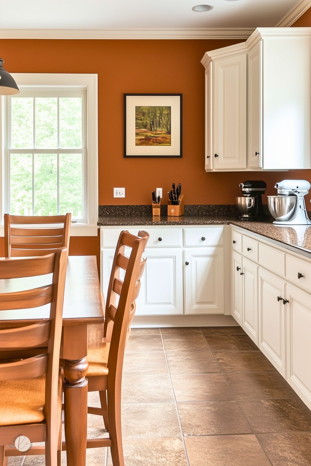 Warm cozy kitchen with burnt-orange walls, white shaker cabinets, dark granite countertops, and brown tile floor. Wood dining table with ladder-back chairs sits by a bright window. Framed landscape art, knife blocks, and stand mixers style the counter. Fall color palette, traditional kitchen design. Text: “Kitchen Informant.”