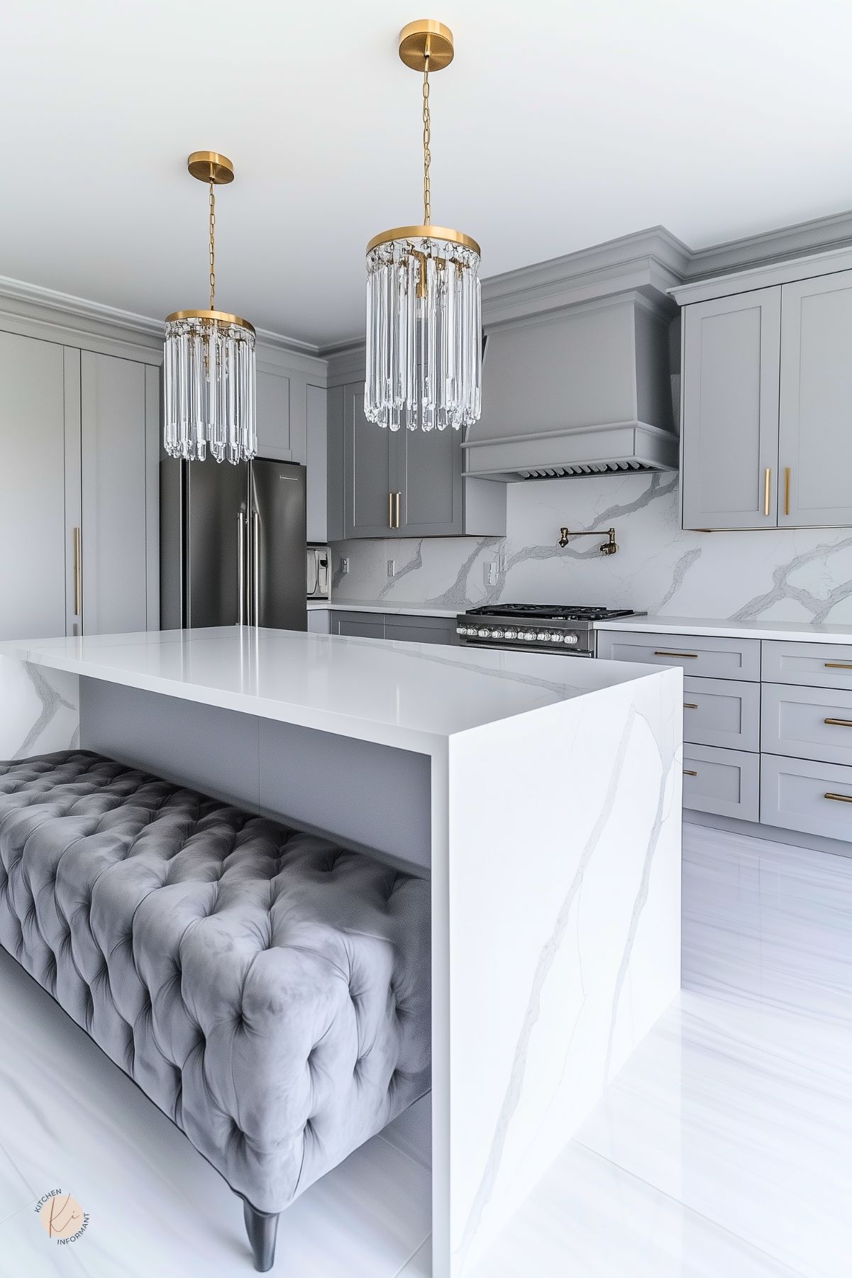 Glam gray and white kitchen with marble waterfall island and tufted gray bench, two gold crystal pendant lights, and light gray shaker cabinets with brass pulls. Coordinating range hood, pot filler, stainless fridge, and veined marble backsplash.