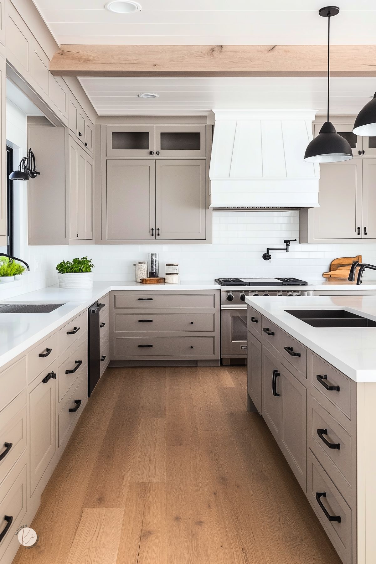 Modern farmhouse kitchen with greige Shaker cabinets and black hardware, white quartz countertops, large island with sink, and white subway tile. White paneled range hood, black pot filler, matte black pendants, induction cooktop, and stainless range. Light oak floors and a white shiplap ceiling with a wood beam. Watermark reads “Kitchen Informant.”