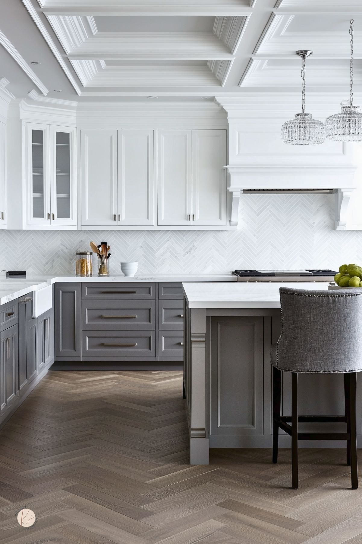 Elegant gray and white kitchen with two-tone cabinets, marble herringbone backsplash, coffered ceiling with heavy molding, and crystal pendants. Large island with white countertop and upholstered counter stool; herringbone wood floors and classic range hood.