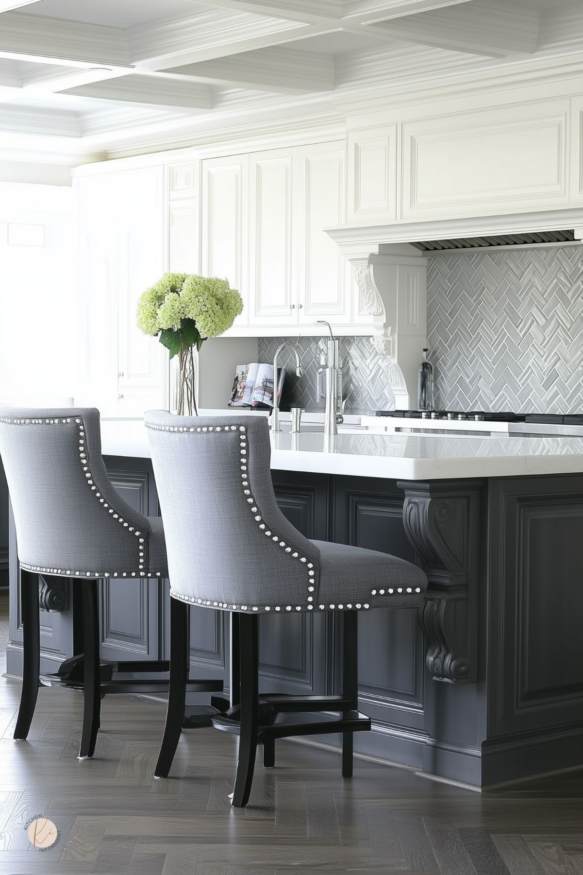 Elegant gray and white kitchen with a coffered ceiling, ornate charcoal island topped with white quartz, and stainless faucet. Two gray nailhead barstools, green hydrangeas in a vase, and a silver herringbone tile backsplash beneath a mantel-style range hood. Herringbone wood floor.