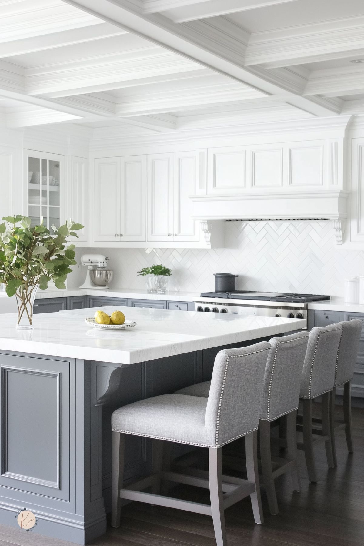 Bright gray and white kitchen with a large marble island, paneled gray base, and three upholstered nailhead barstools. White shaker cabinets, coffered ceiling, and a white herringbone tile backsplash beneath a mantel range hood. Vase with greenery and a plate of lemons.