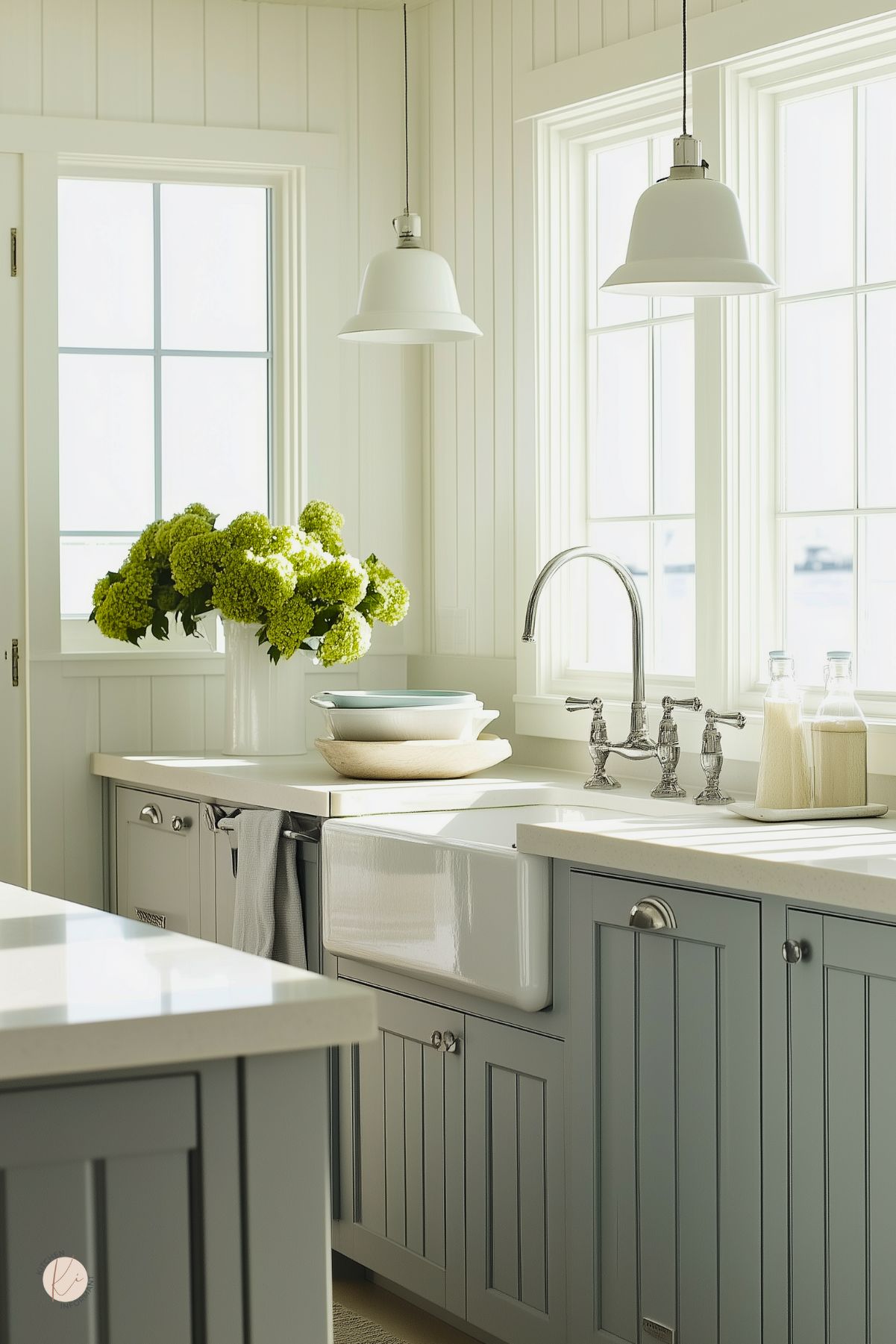 Bright coastal kitchen with light gray shaker cabinets and white quartz counters. Apron-front farmhouse sink with bridge faucet beneath two white pendants and tall windows. Vase of green hydrangeas, stacked bowls, and bottles on the counter.