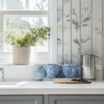 Light-filled cottage kitchen with grey shaker cabinets and white quartz countertop. Floral bird wallpaper and a white Roman shade around the window. Potted greenery on the sill, chrome faucet, blue and white ceramic jugs/mugs, a round wood board with knife, and a stainless canister. Subtle “KI” logo in the corner. Keywords: cottage kitchen, grey cabinets, floral wallpaper.