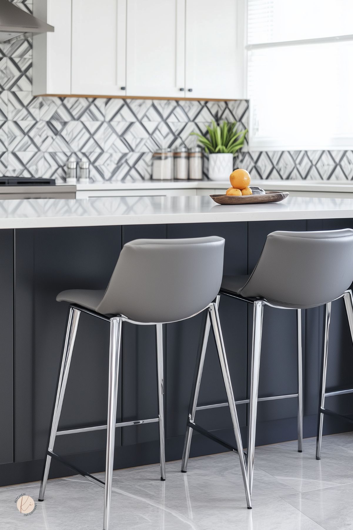 Modern grey and white kitchen with a charcoal island and white quartz countertop. Two gray bar stools with chrome legs face a geometric black-and-white tile backsplash. White uppers, stainless range hood, canisters and a potted plant along the counter; wooden tray with citrus. Small “KI” logo in corner.