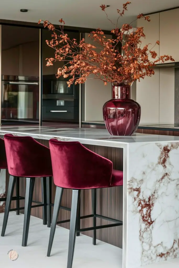Modern kitchen design with burgundy accents: velvet counter stools and a large glossy burgundy vase with dried branches atop a marble waterfall island with brown veining. Sleek slab-front cabinetry, integrated black appliances, and wood paneling complete the contemporary look.