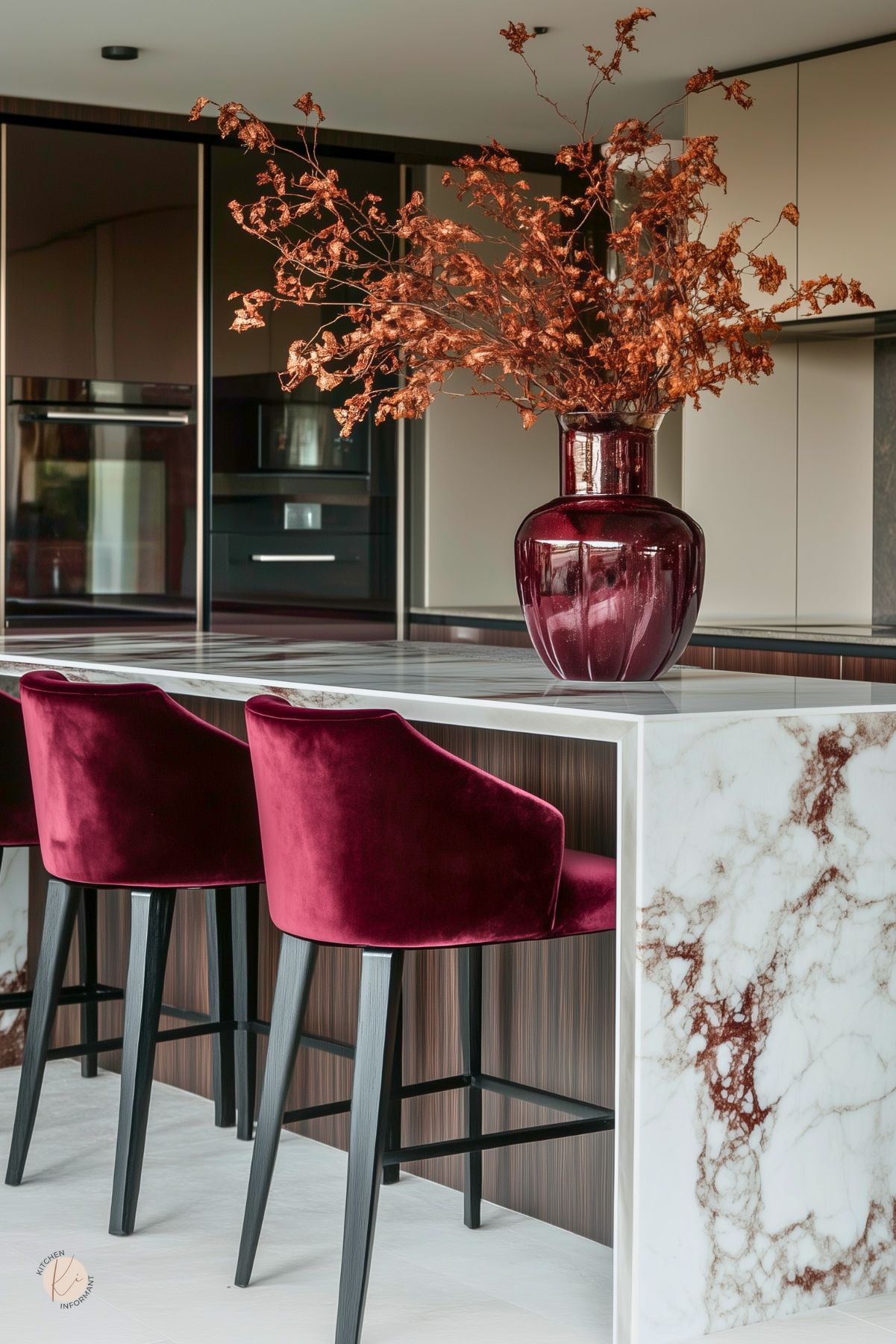 Modern kitchen design with burgundy accents: velvet counter stools and a large glossy burgundy vase with dried branches atop a marble waterfall island with brown veining. Sleek slab-front cabinetry, integrated black appliances, and wood paneling complete the contemporary look.