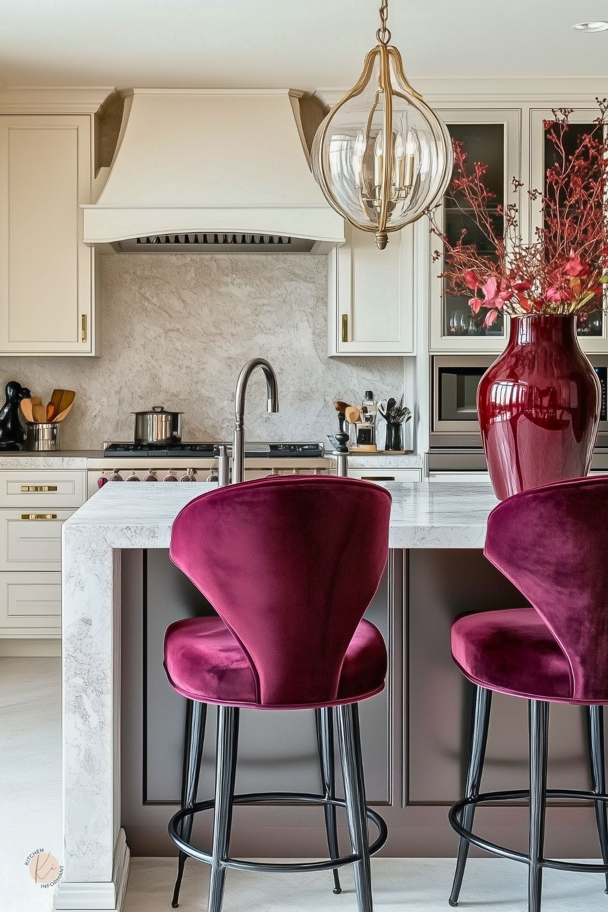 Elegant kitchen design with white shaker cabinets, plaster range hood, and marble waterfall island. Burgundy accents: velvet bar stools and a glossy vase with branches. Brass hardware, glass globe pendant light, and stone-look backsplash complete the look. Marble island, burgundy kitchen decor.