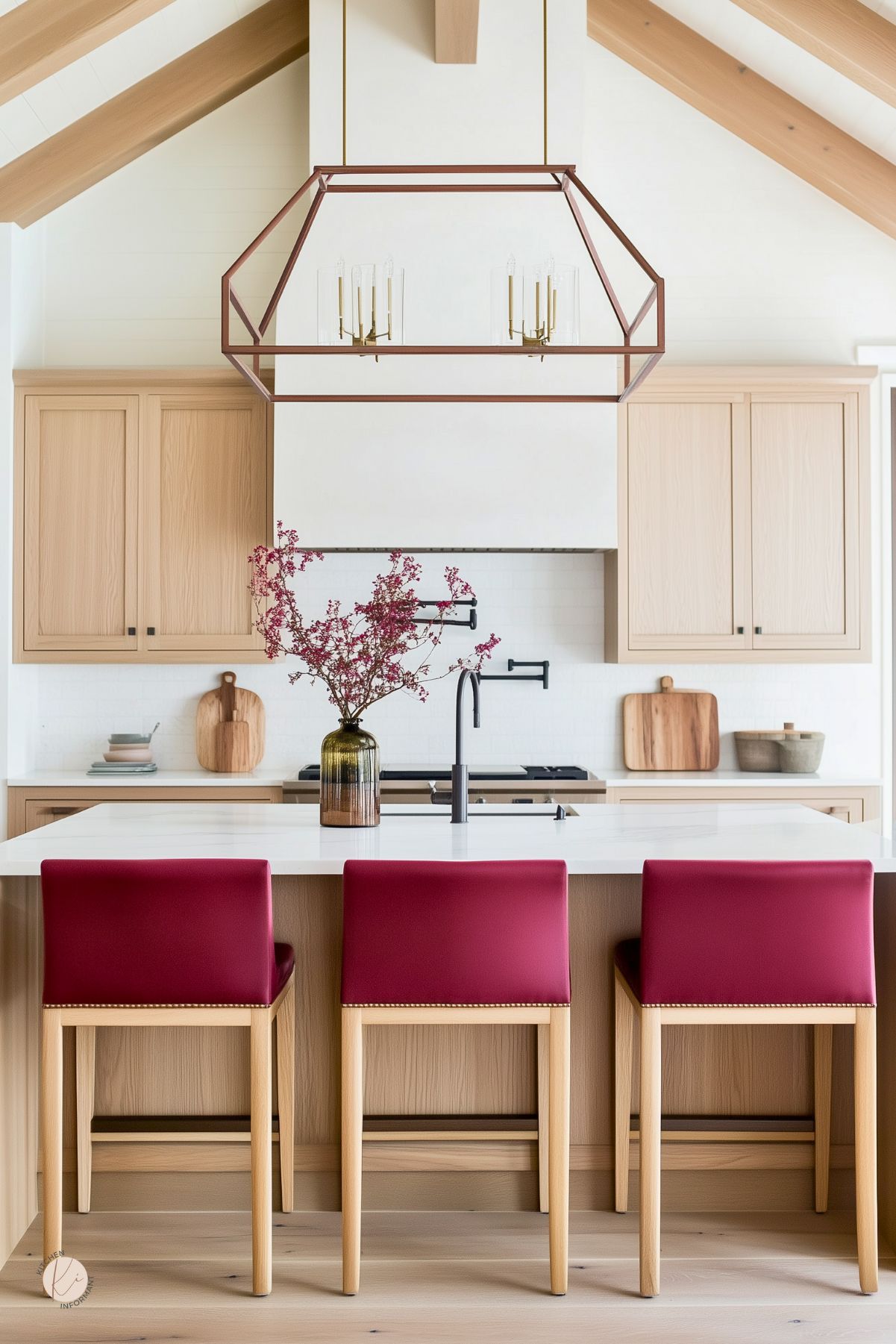 Airy kitchen design with white oak cabinets and beams, a large geometric pendant, and a white quartz island. Three burgundy leather bar stools with nailhead trim face a black gooseneck faucet. White plaster hood, dual pot fillers, and a vase with pink branches add soft accents.