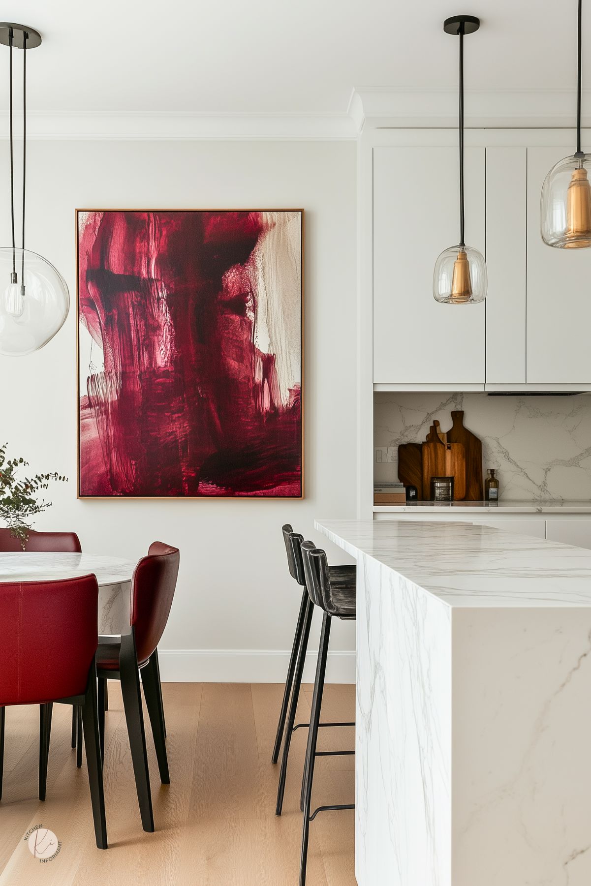Modern kitchen and dining area with burgundy accent chairs around a white marble dining table. A bold abstract burgundy painting hangs on the wall. The space includes a white waterfall-edge marble island with black barstools, glass pendant lights, light wood flooring, and minimalist white cabinetry.