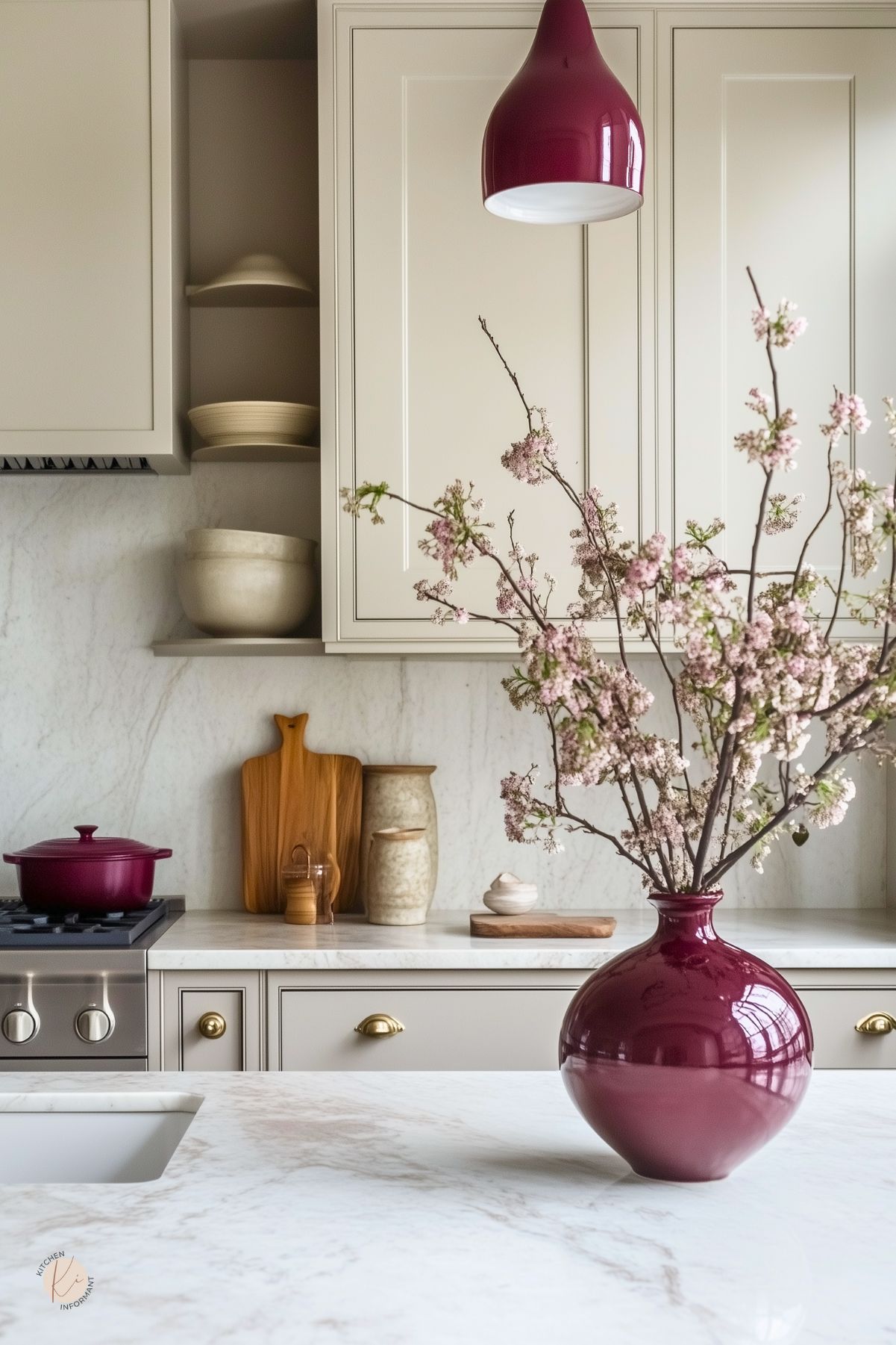 Warm greige kitchen with marble countertops and slab backsplash, shaker cabinets, and brass hardware. Burgundy accents: glossy pendant light, round vase with flowering branches, and a Dutch oven on the range. Styled with wood cutting boards and neutral pottery. Kitchen design.