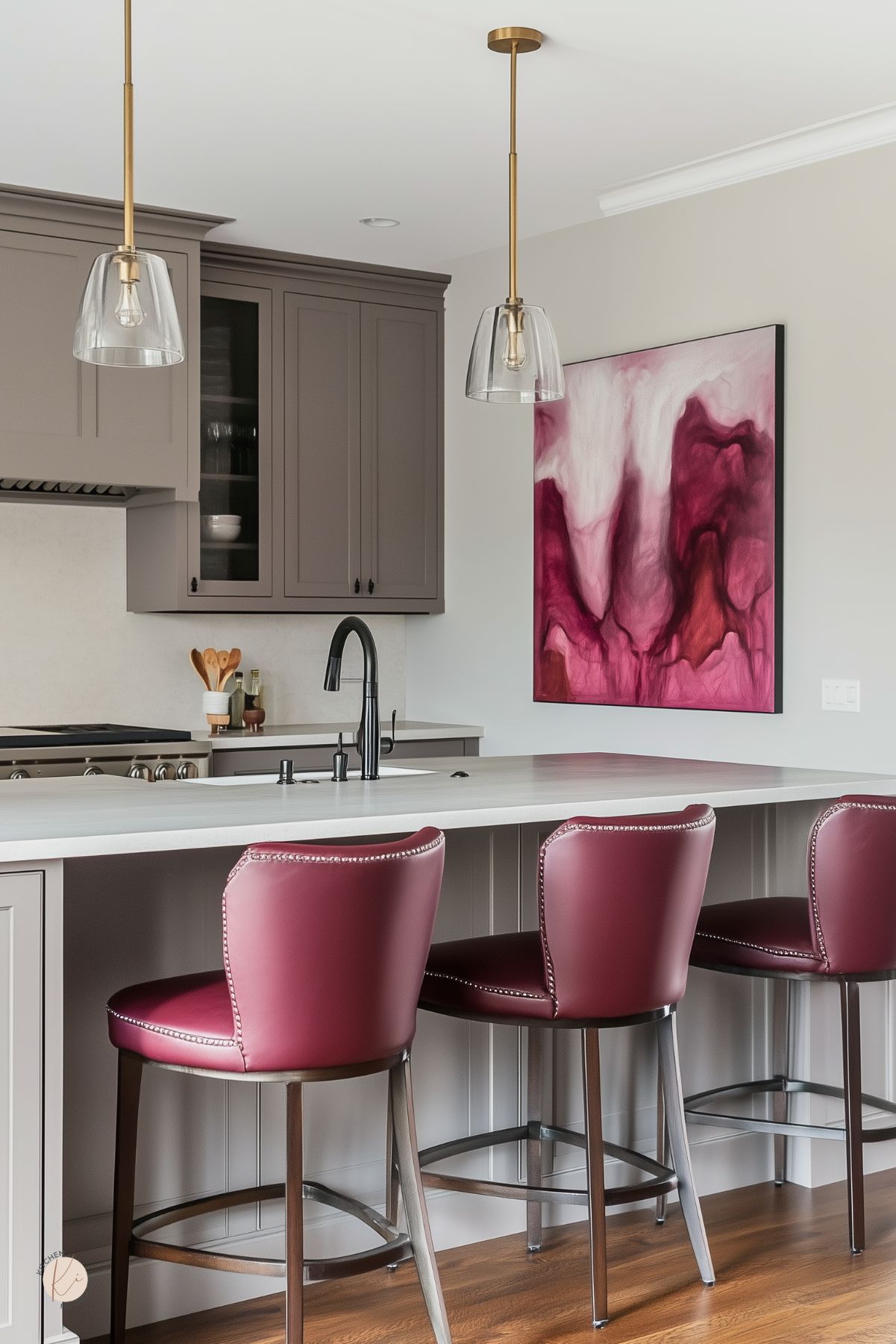 Taupe kitchen with a large light-quartz island, black pull-down faucet, and brass-and-glass pendant lights. Three burgundy leather bar stools with nailhead trim line the island. Shaker cabinets and wood floors, plus a large abstract burgundy art piece add rich kitchen design accents.