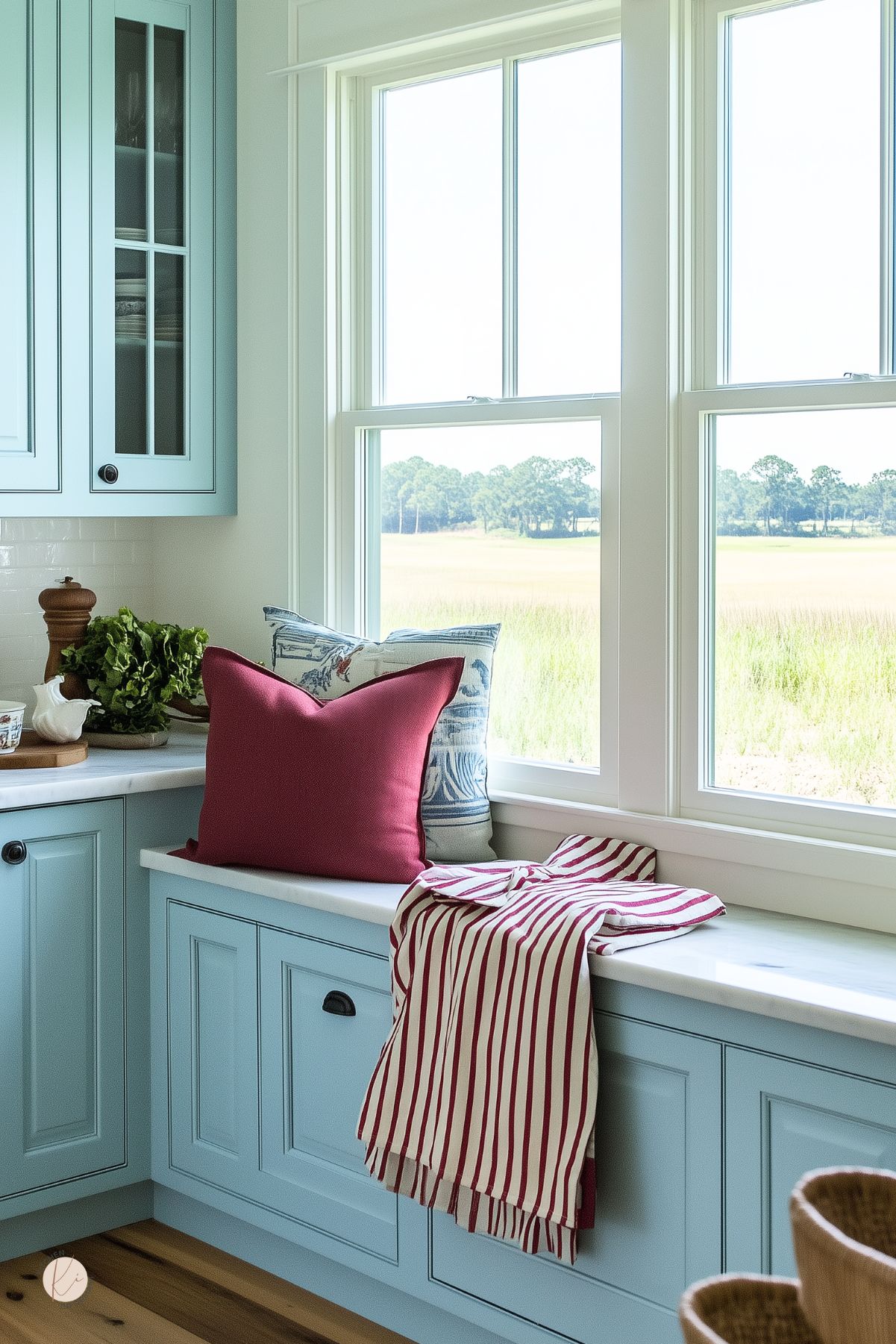 Coastal-style kitchen with soft blue cabinetry and a bright window seat. A solid burgundy pillow and a red-and-white striped throw add bold contrast against the pale blue and white backdrop. Blue toile pillow and natural wood accents complete the inviting corner.