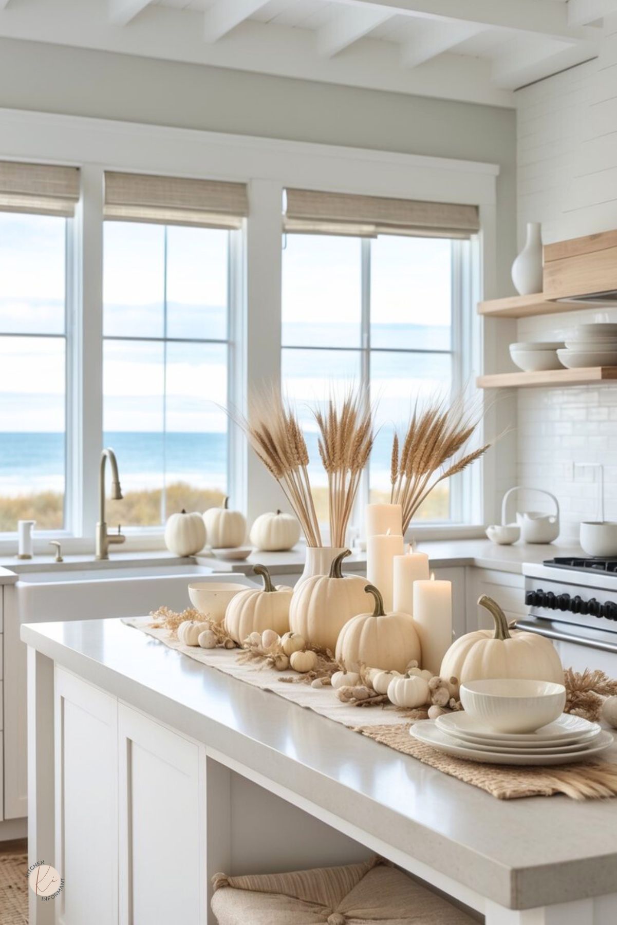 Neutral Thanksgiving kitchen decor in a bright coastal kitchen: white cabinets, farmhouse sink, and ocean view. Island styled with a beige runner, white pumpkins, wheat bundle centerpiece, pillar candles, and stacked white dishes; open wood shelves and range at right. Minimal, neutral fall kitchen styling. Small Kitchen Informant logo.