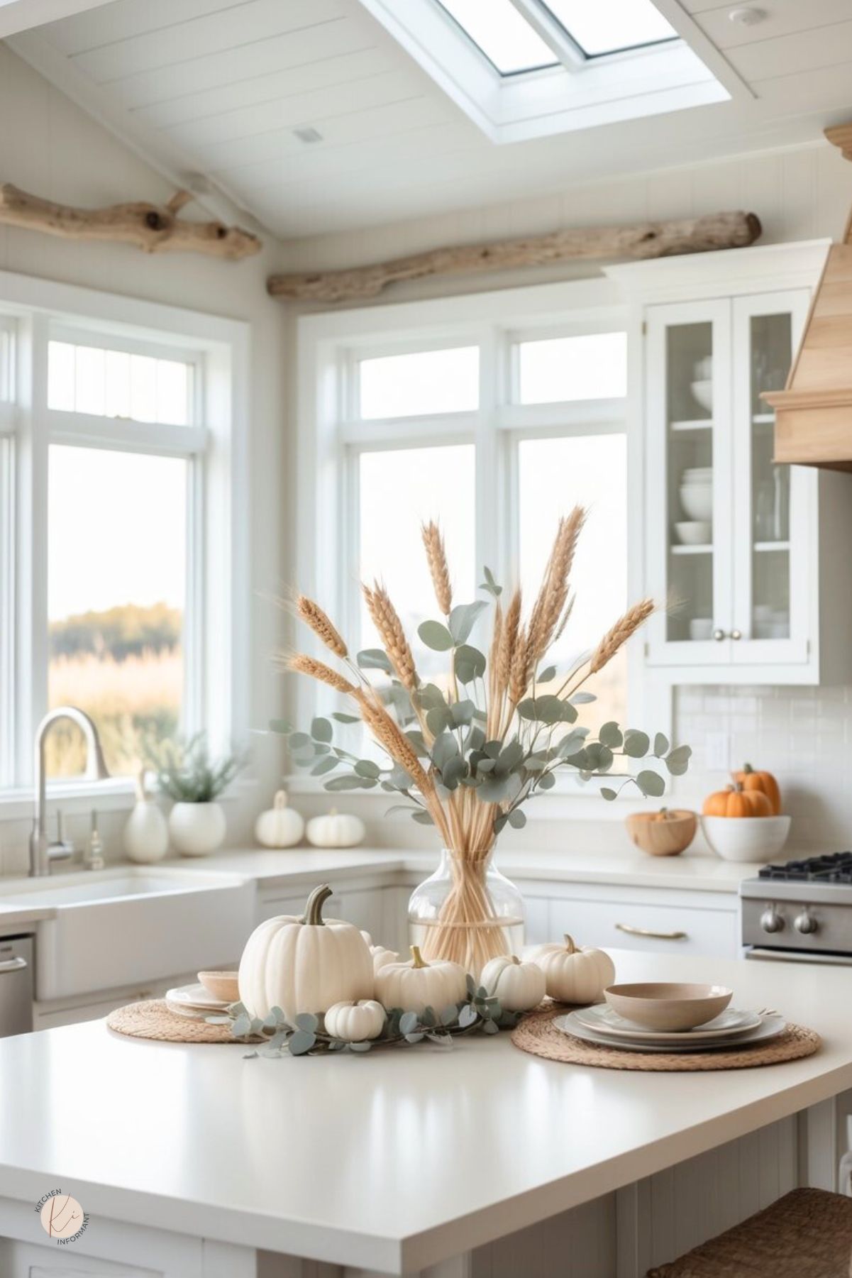 Neutral Thanksgiving kitchen decor: bright white kitchen with skylight, farmhouse sink, and shaker cabinets. Island styled with white pumpkins, eucalyptus garland, and a glass vase of wheat; woven placemats with stacked dishes. Warm wood accents and window views. Small Kitchen Informant logo.
