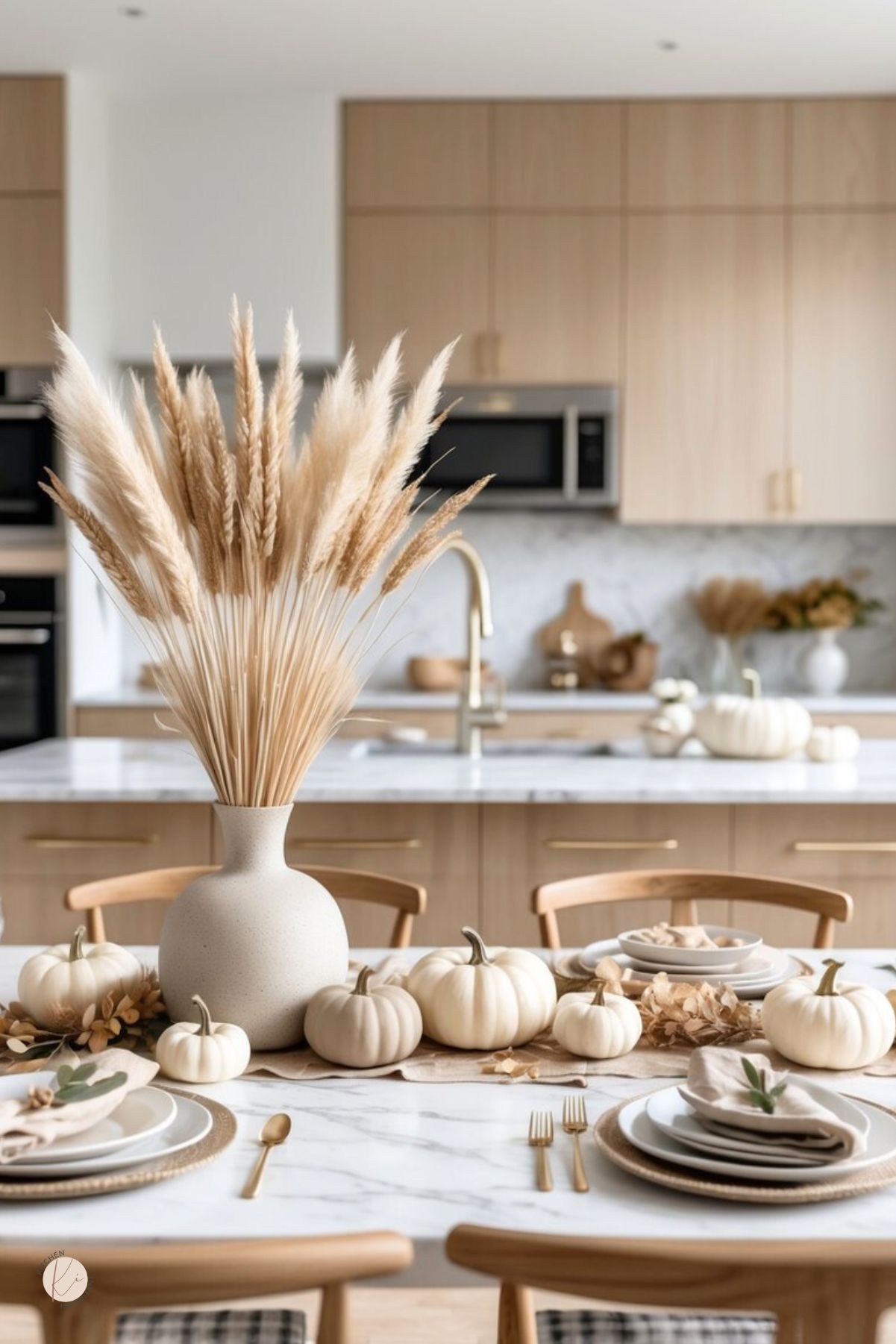 Neutral Thanksgiving kitchen tablescape with a pampas-grass centerpiece, white pumpkins lining a marble table, linen runner, layered plates with linen napkins, and gold flatware. Modern light wood cabinets, marble island, and brass faucet in back. Neutral fall kitchen decor, Thanksgiving table setting. Small Kitchen Informant logo.