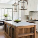 Modern farmhouse kitchen with white shaker cabinets, marble subway tile backsplash, and a rustic wood island topped with white marble. Two large lantern pendant lights, wide black-frame windows, wood floors, and a bowl of green apples on the island; dining nook in back. Small “KI” logo in corner.
