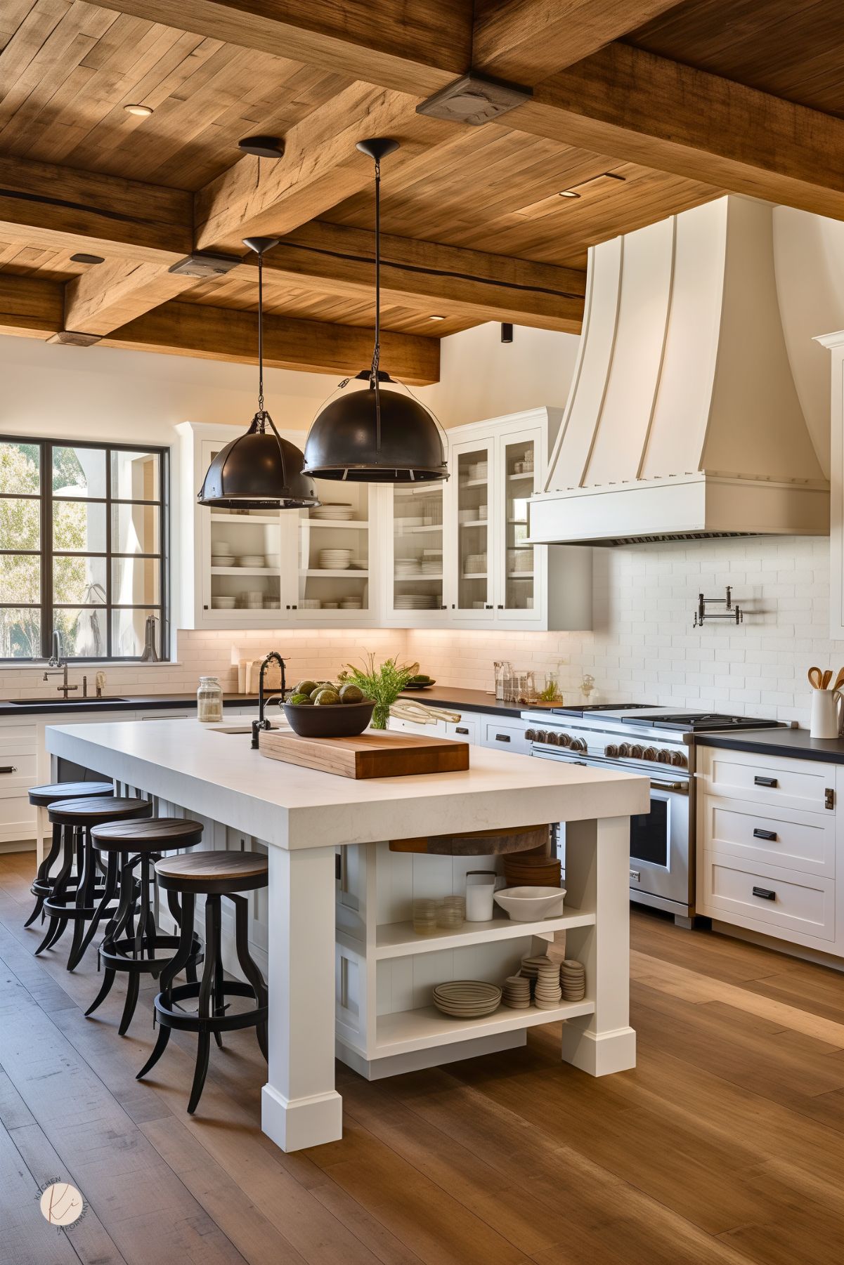 Rustic farmhouse kitchen with wood-beam ceiling, white shaker cabinets and glass-front uppers, black countertops, and a large white quartz island with open shelves and turned stools. Oversize black dome pendants, statement range hood with pot filler, grid windows, and warm wood floors. Small “KI” logo in corner.