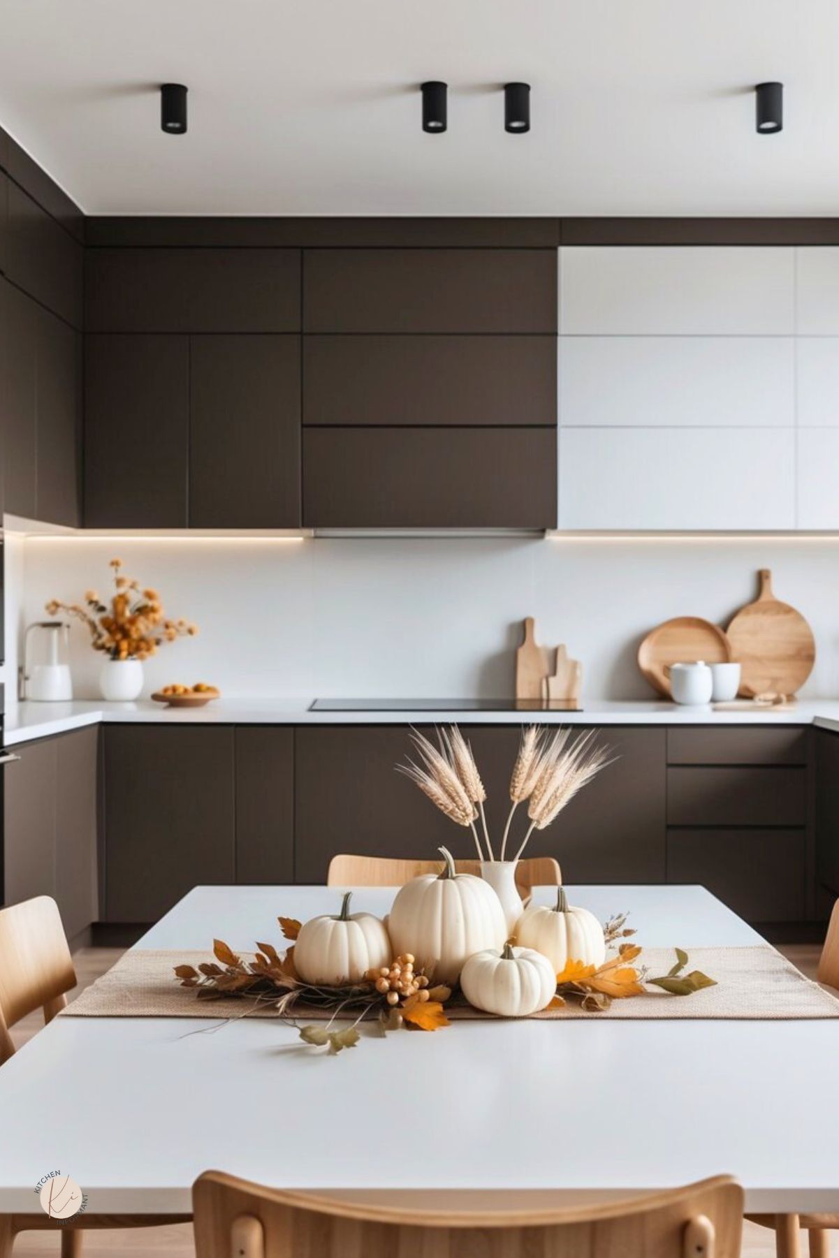 Modern minimalist kitchen with flat-panel dark brown and white cabinets, white countertops, and wood accents. A white dining table holds a burlap runner and simple Thanksgiving centerpiece: white pumpkins, wheat stems, and fall leaves. Neutral fall kitchen decor. Text: “Kitchen Informant.”