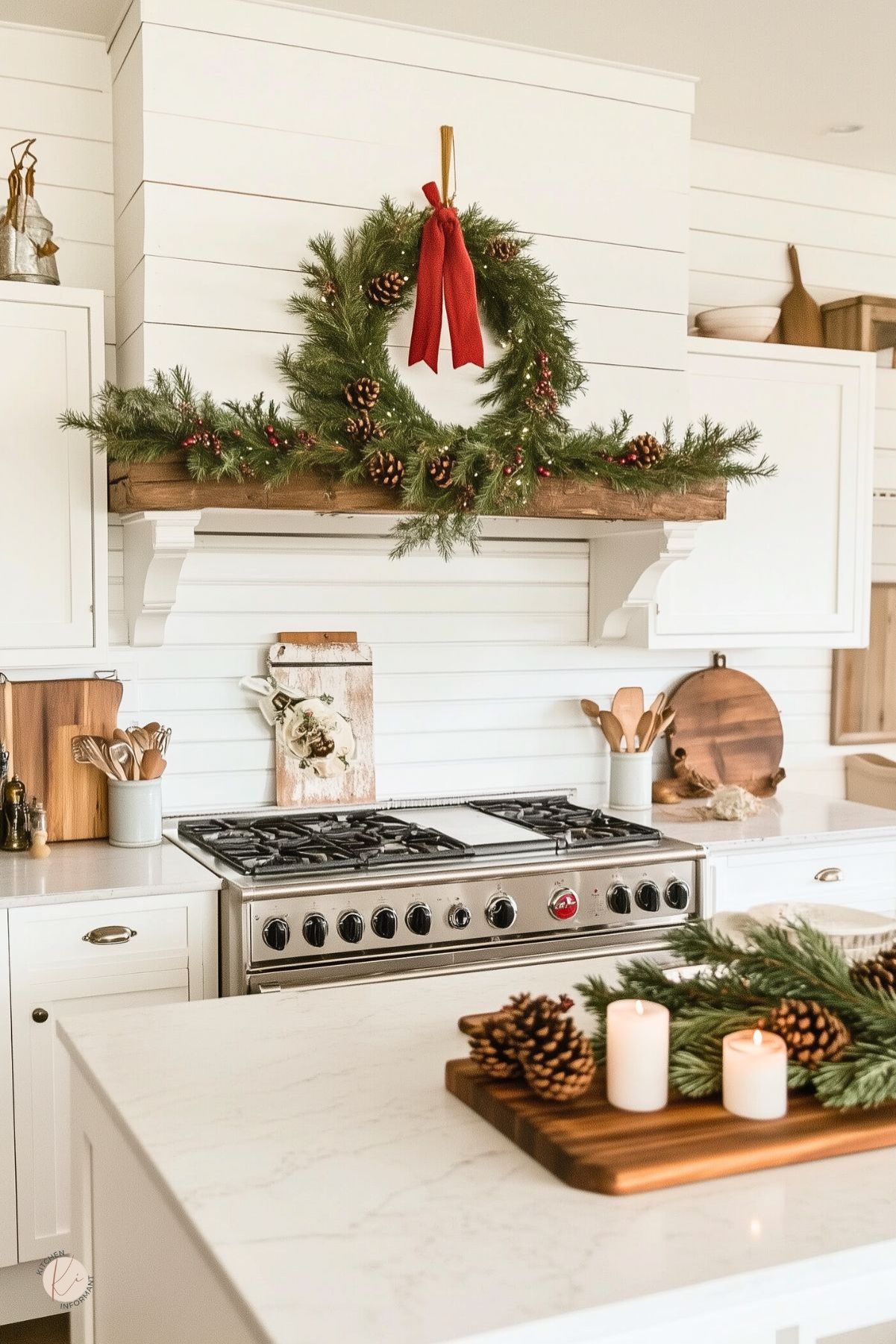 Bright white farmhouse kitchen decorated for Christmas with a green wreath and red ribbon above the stove, pine garland on a wooden shelf, and warm neutral decor. A large stainless steel range, white cabinets, cutting boards, and a kitchen island styled with candles, pinecones, and greenery create a cozy holiday kitchen look.