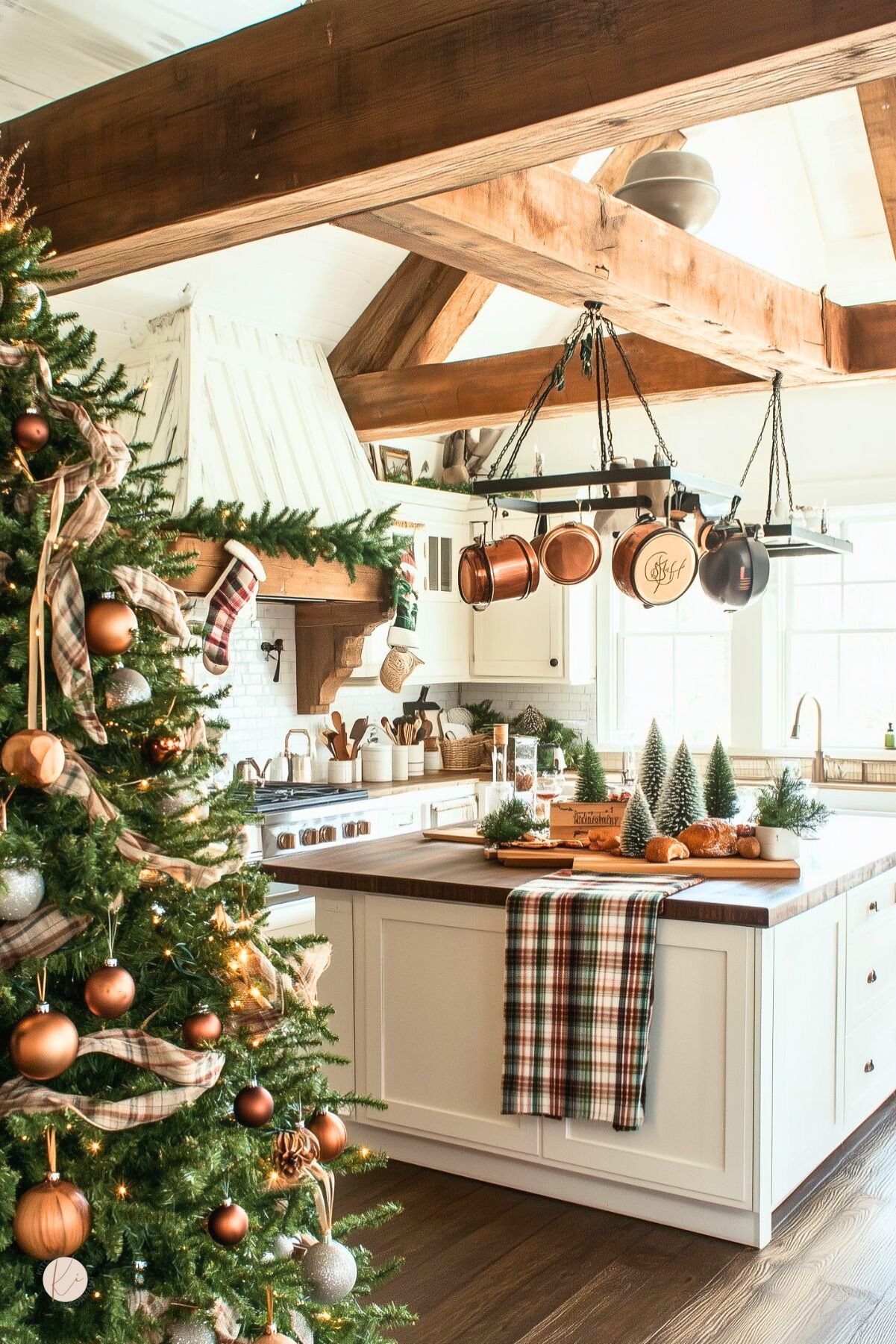 Rustic farmhouse kitchen decorated for Christmas with exposed wood beams, white cabinets, and a large island topped with greenery and mini trees. A decorated Christmas tree with plaid ribbon and copper ornaments stands beside the island. Hanging copper pots, garland, stockings, and warm natural light create a cozy holiday kitchen.