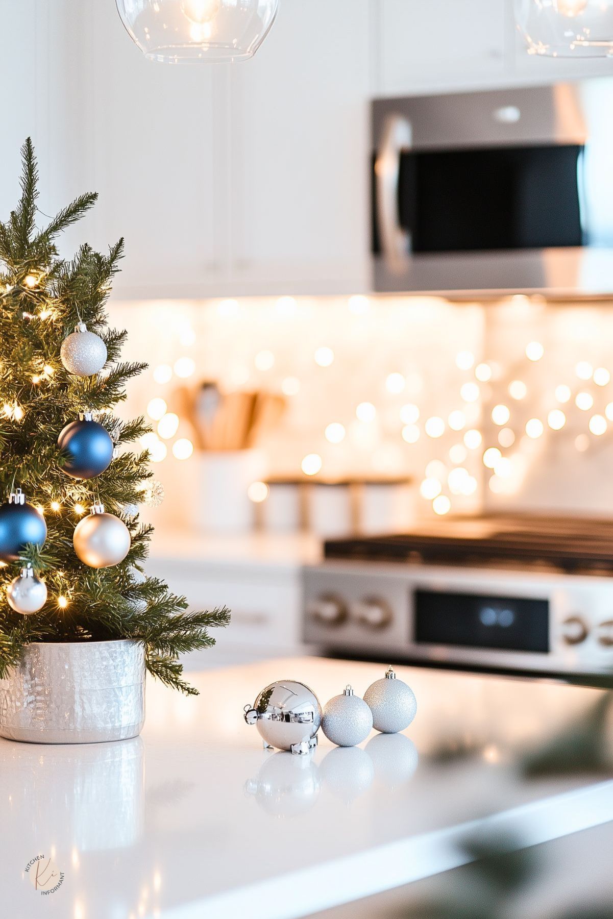 Minimalist white kitchen decorated for Christmas with a small tabletop tree in a silver pot, blue and silver ornaments, and warm string lights along the backsplash. A white quartz island holds silver baubles, with stainless steel appliances and soft pendant lighting creating a clean, modern holiday kitchen look.