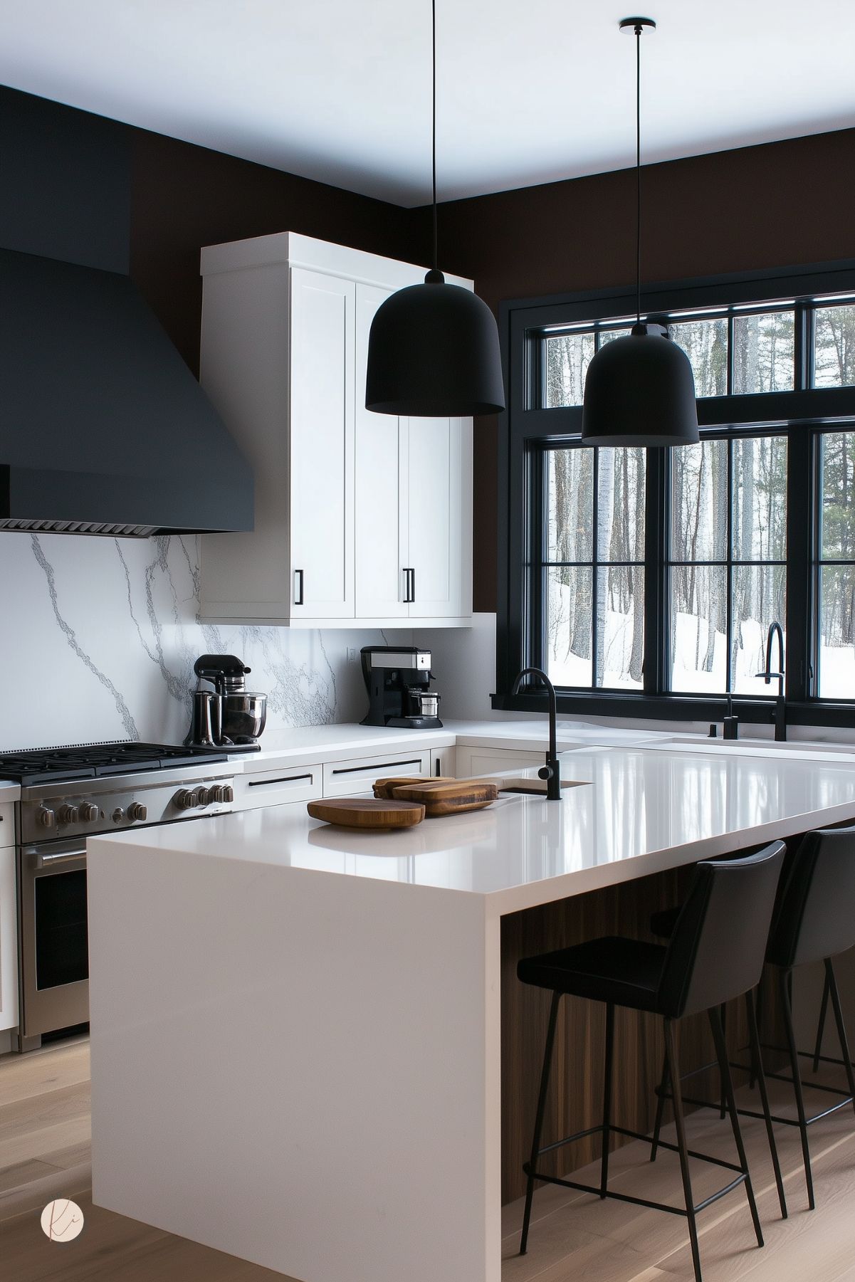 Brown and white kitchen with a sleek white waterfall island, black pendant lights, and black-framed windows overlooking snowy trees. White cabinets and marble-look backsplash pair with a black range hood, while dark wood paneling under the island and black bar stools add warm contrast to the modern kitchen design.