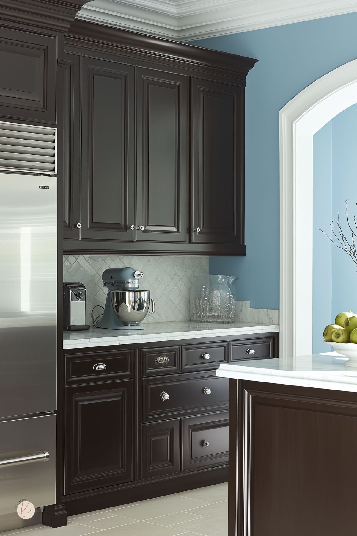 Dark brown traditional kitchen with tall paneled cabinets, white marble countertops, and a pale herringbone tile backsplash. Soft blue walls and a white arched doorway brighten the space, while stainless appliances, chrome hardware, a blue stand mixer, and a bowl of green apples add polished detail.
