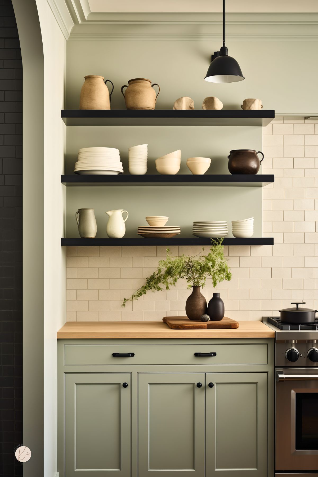 Sage green kitchen wall with black floating shelves, cream subway tile backsplash, butcher block countertop, and black hardware. Neutral pottery, stacked plates, and a small vase with airy greenery style the shelves, while a black pendant light hangs above. Edge of a stainless range shows on the right. Faint “Kitchen Informant” watermark at bottom left.