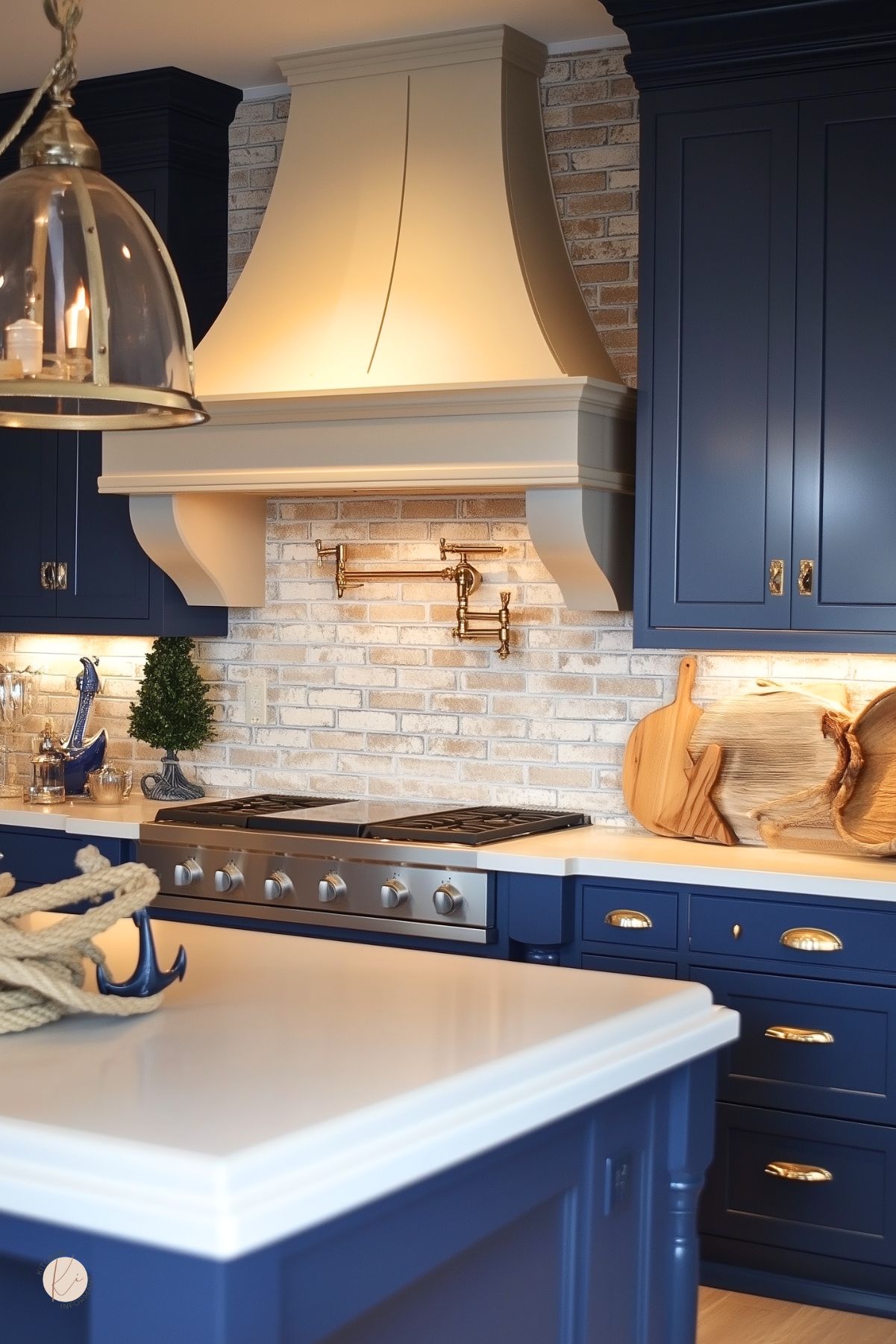 Navy blue kitchen with a white brick backsplash, white range hood, brass pot filler, and white countertops. Stainless steel range sits between dark blue shaker cabinets with brass hardware. Large white island in front with a rope and blue anchor decor piece. Warm under-cabinet lighting and wood cutting boards add coastal kitchen design style.