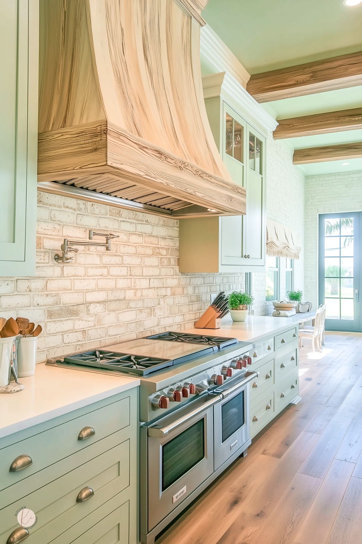 Light sage green kitchen with a white brick backsplash, oversized natural wood range hood, and matching exposed ceiling beams. Long white countertop with a large stainless steel range and red knobs, pot filler, pale wood floors, and soft farmhouse kitchen design details leading to a bright dining nook.