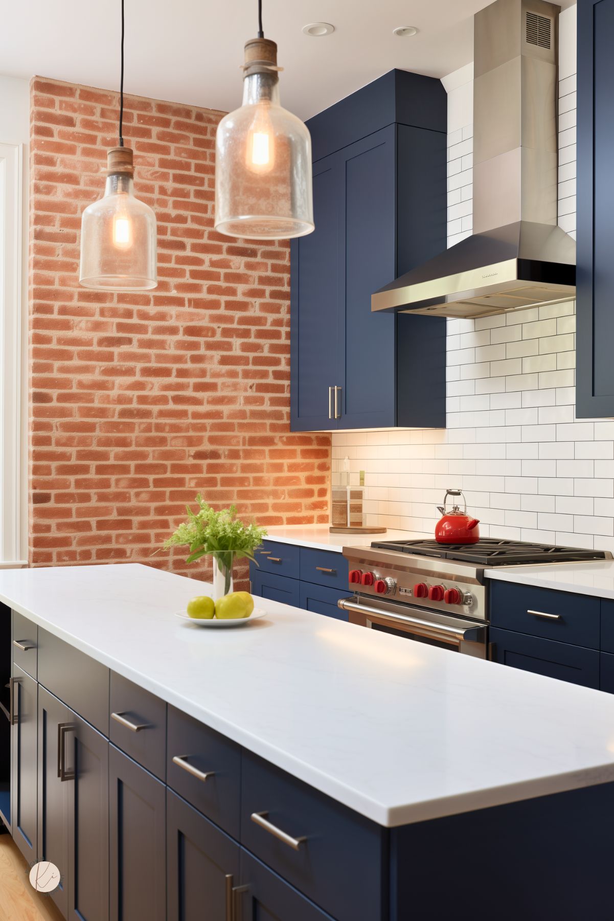 Navy blue kitchen with a red brick accent wall, white subway tile backsplash, and large white island. Stainless steel range with red knobs sits below a sleek hood, while glass pendant lights hang overhead. White countertops, silver hardware, green apples, and a red kettle add contrast.