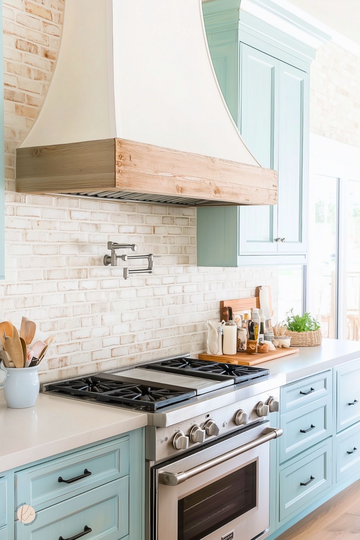 Light blue kitchen with a white painted brick backsplash, oversized white range hood with natural wood trim, and stainless steel range. Pale blue shaker cabinets, white countertops, black hardware, and a wall pot filler create a bright coastal farmhouse kitchen design.
