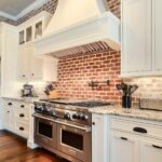 White kitchen with a red brick backsplash, large curved white range hood, and stainless steel double oven range. White raised-panel cabinets, black hardware, marble-look countertops, and dark wood floors create a bright farmhouse kitchen design with classic brick backsplash style.